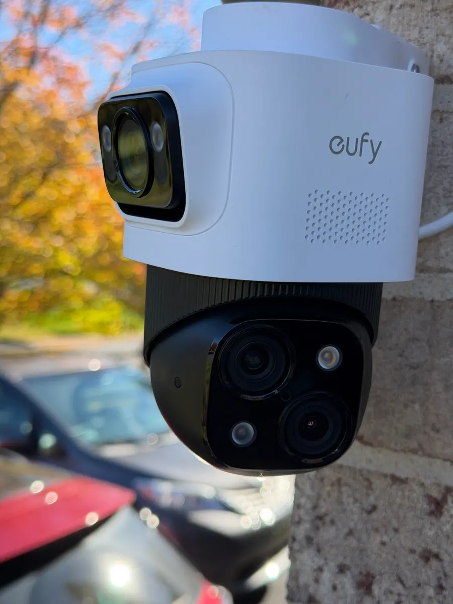 A white dual-lens security camera on a brick wall above parked cars.