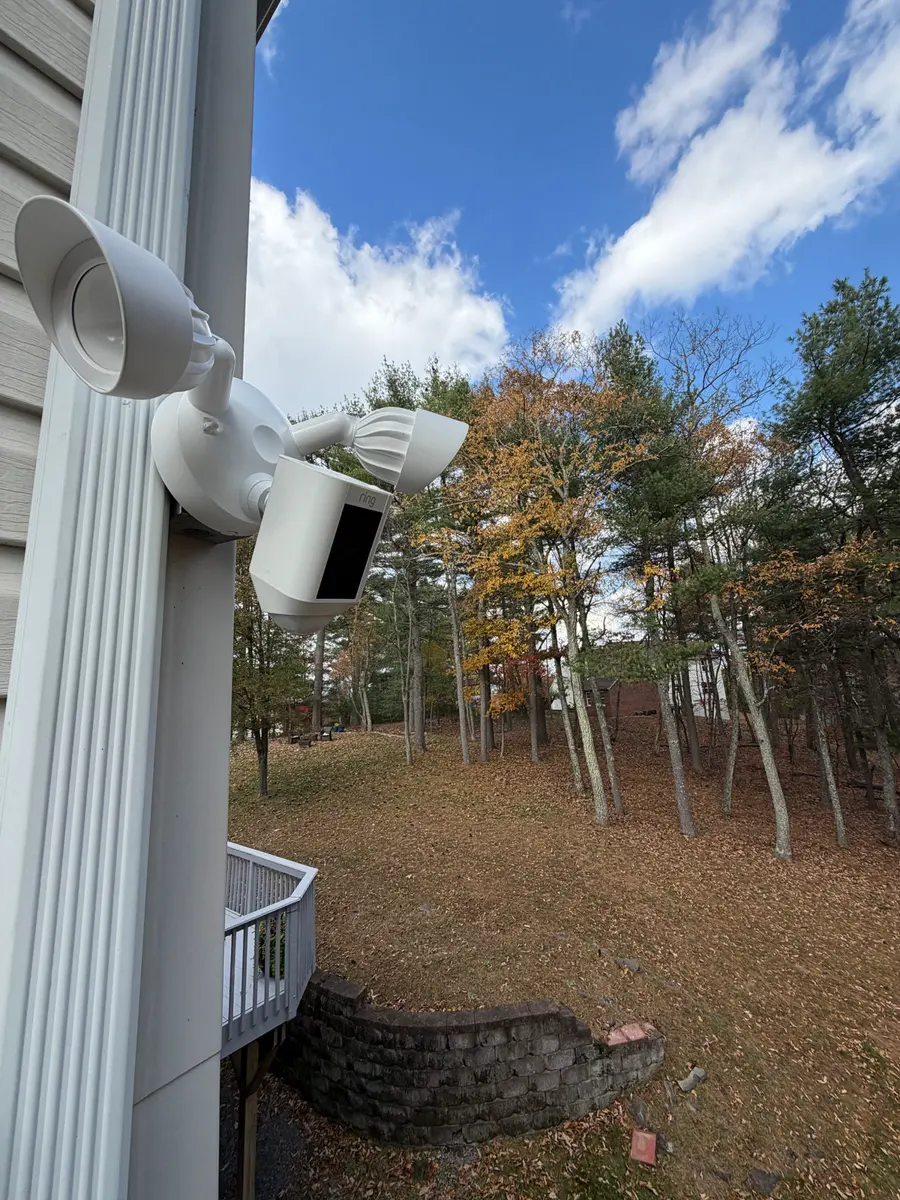 A floodlight camera on the corner of a house overlooking a wooded yard.